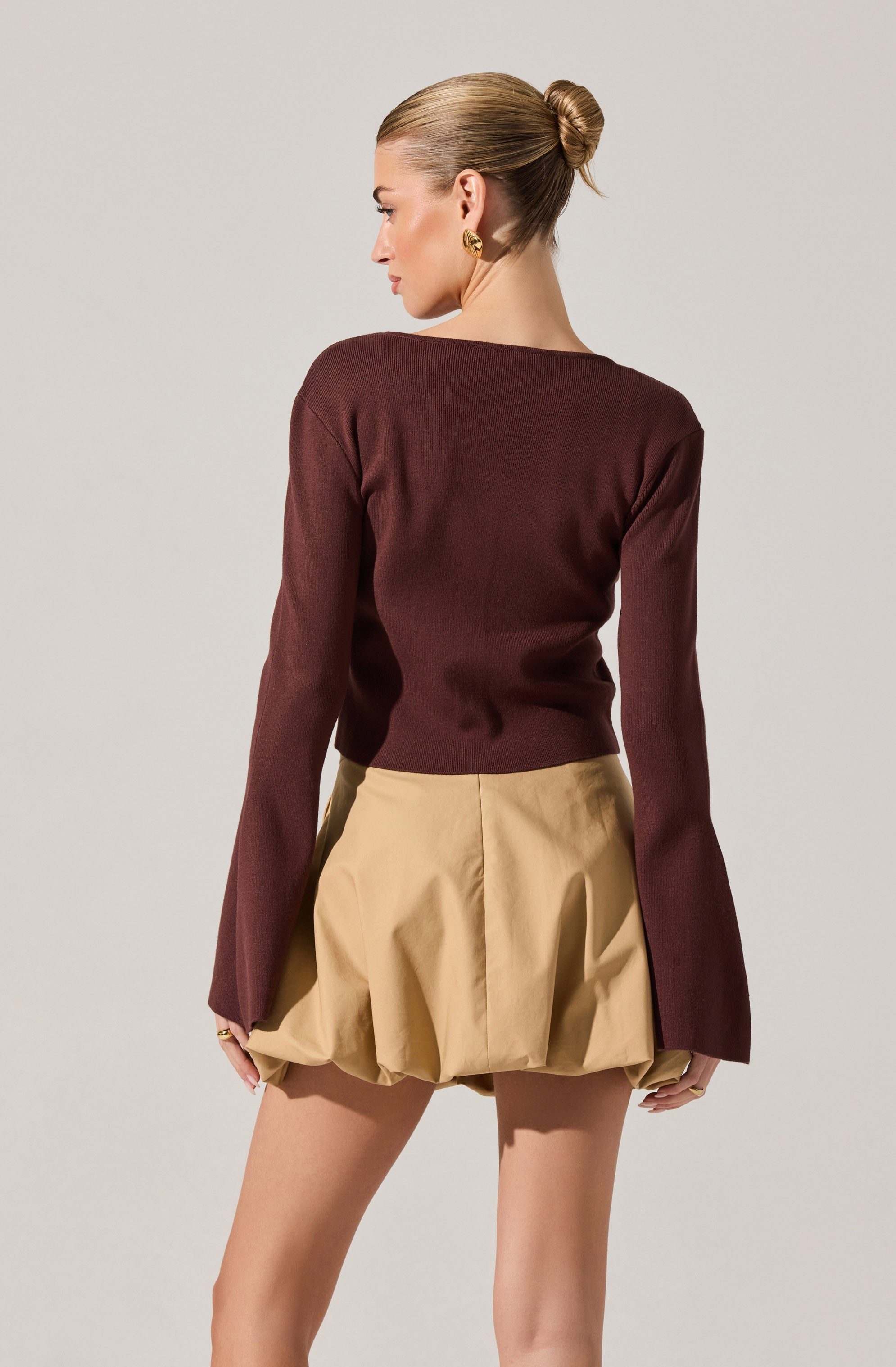 A woman is wearing a long-sleeved, dark red (burgundy) sweater with a round neckline. She is also wearing beige, loose-fitting shorts with pleats, giving a puffed appearance at the hem. The outfit is complemented by gold hoop earrings. The woman has her hair styled in a sleek, tight bun and is facing away from the camera, showing the back of her clothing.