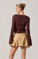 A woman is wearing a long-sleeved, dark red (burgundy) sweater with a round neckline. She is also wearing beige, loose-fitting shorts with pleats, giving a puffed appearance at the hem. The outfit is complemented by gold hoop earrings. The woman has her hair styled in a sleek, tight bun and is facing away from the camera, showing the back of her clothing.