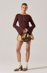 A woman is wearing a fitted, long-sleeved, maroon cardigan with a row of gold buttons down the front and flared sleeves. She has paired it with beige high-waisted shorts, cream-colored socks, and brown high-heeled shoes with bow details. She is carrying a small dark brown handbag with a gold buckle.