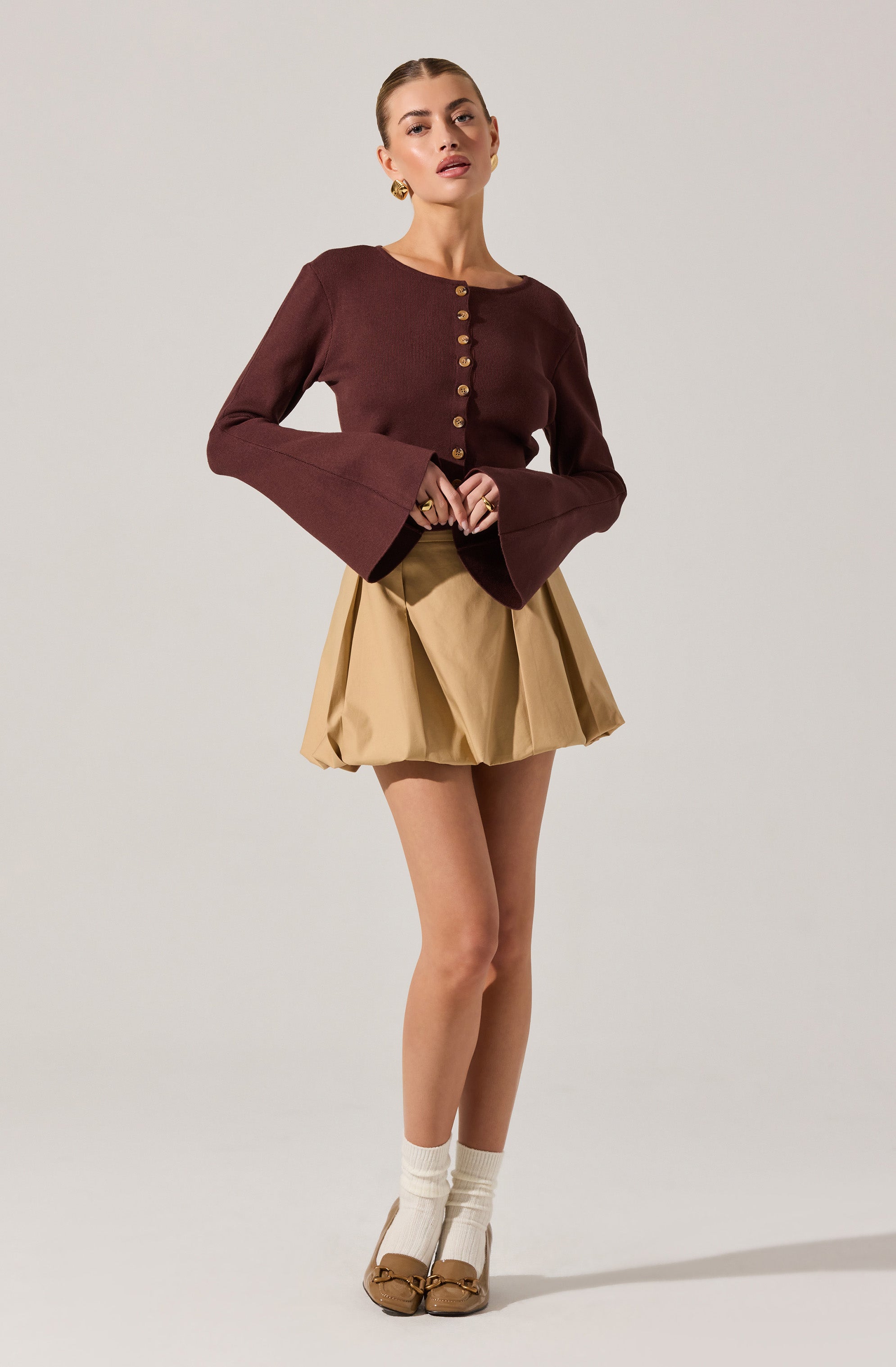 A woman is wearing a long-sleeved, dark brown cardigan with gold buttons down the front, fitted at the waist with bell-shaped sleeves. She pairs it with a high-waisted, beige, pleated, puffy mini skirt. She is also wearing white socks and beige ballet flats with bow details.