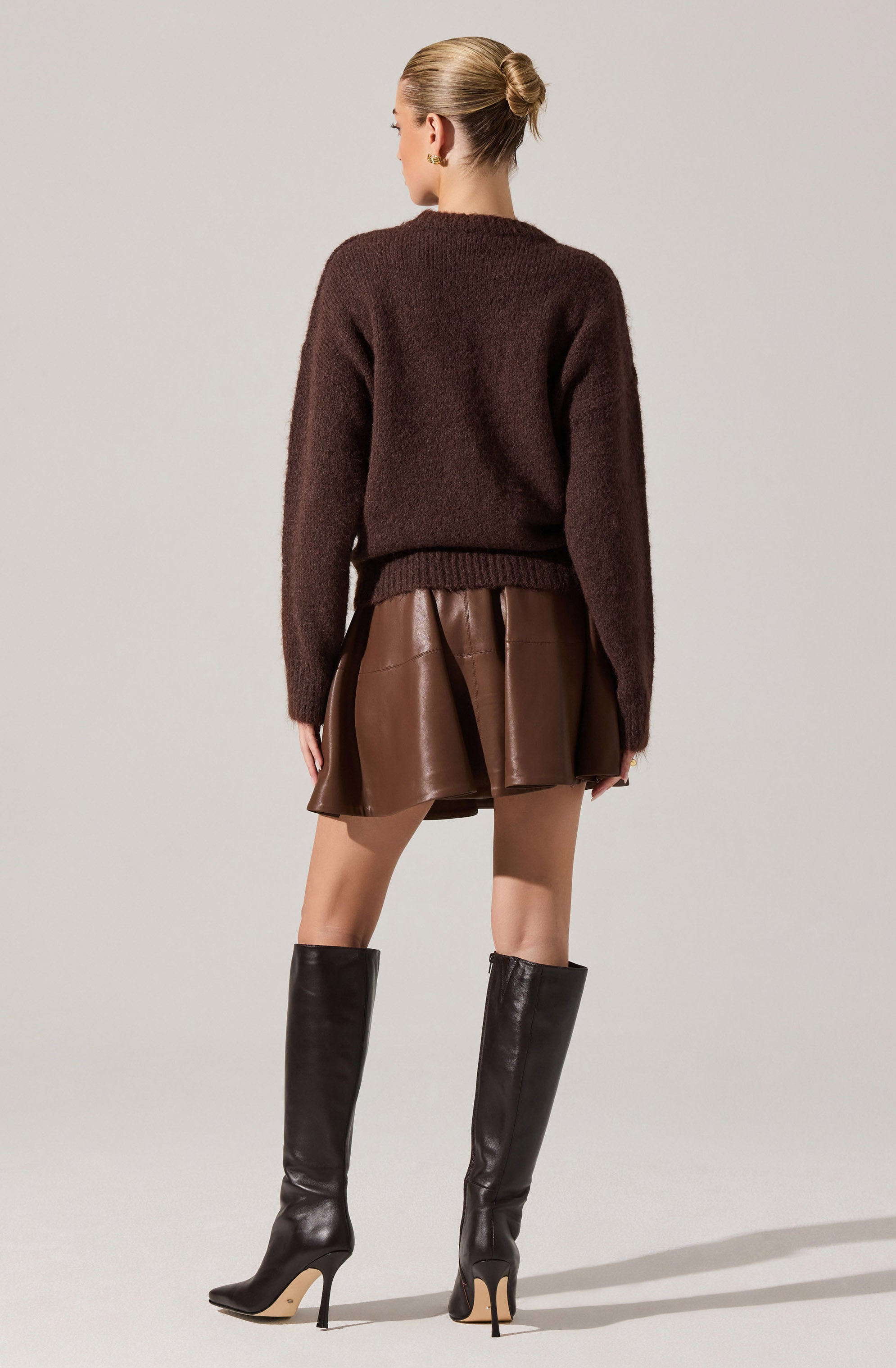 A woman is wearing a loose, dark brown knit sweater with a ribbed hem and cuffs. She pairs it with a shiny, brown leather mini skirt that has pleats. She completes her outfit with tall, black leather high-heeled boots.