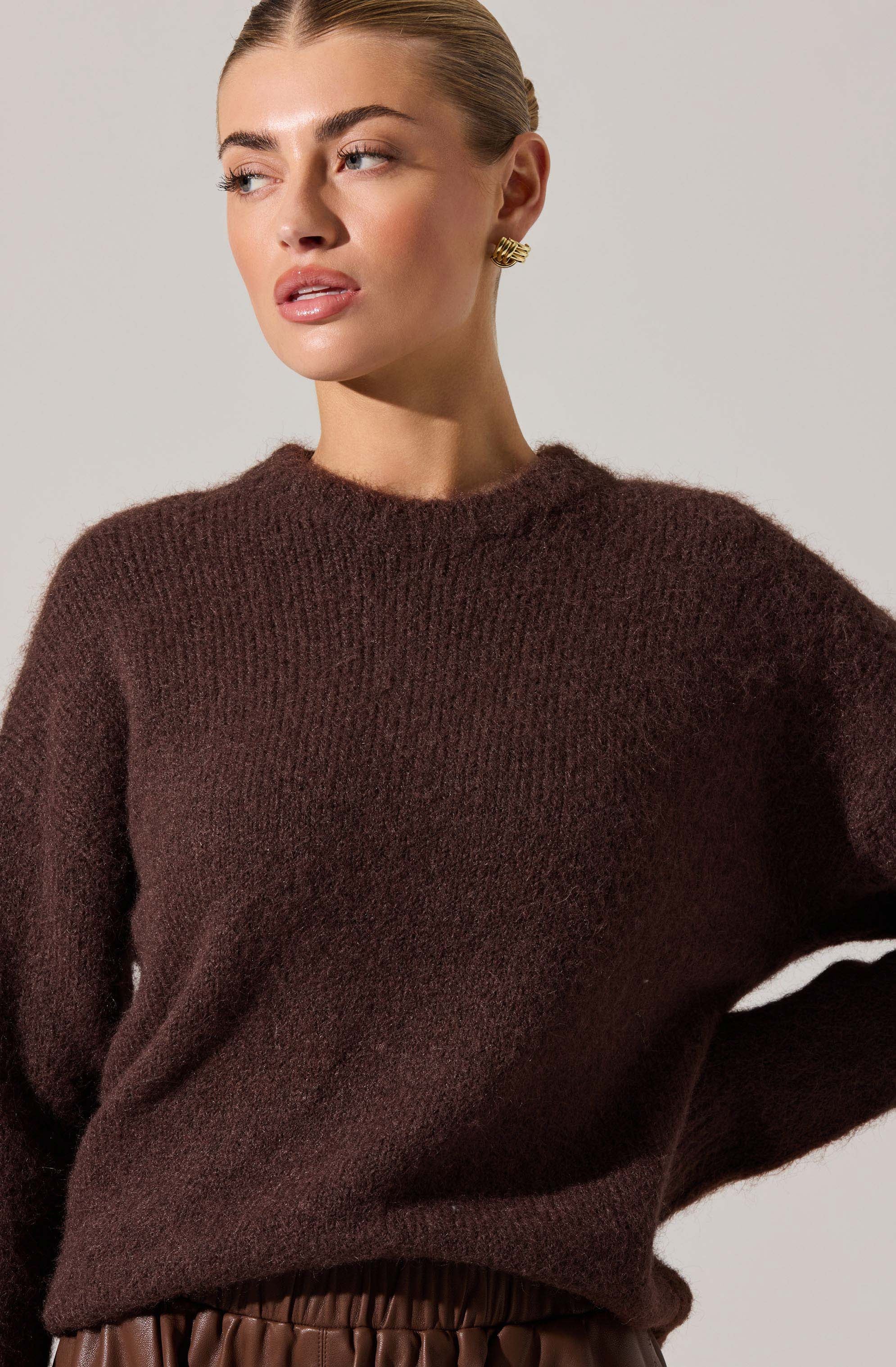 A woman is wearing a dark brown, fuzzy knit sweater with a round neckline. She has accessorized with a pair of brooch-style earrings. The sweater appears soft and slightly oversized, and she has tucked part of the sweater into her waistband.