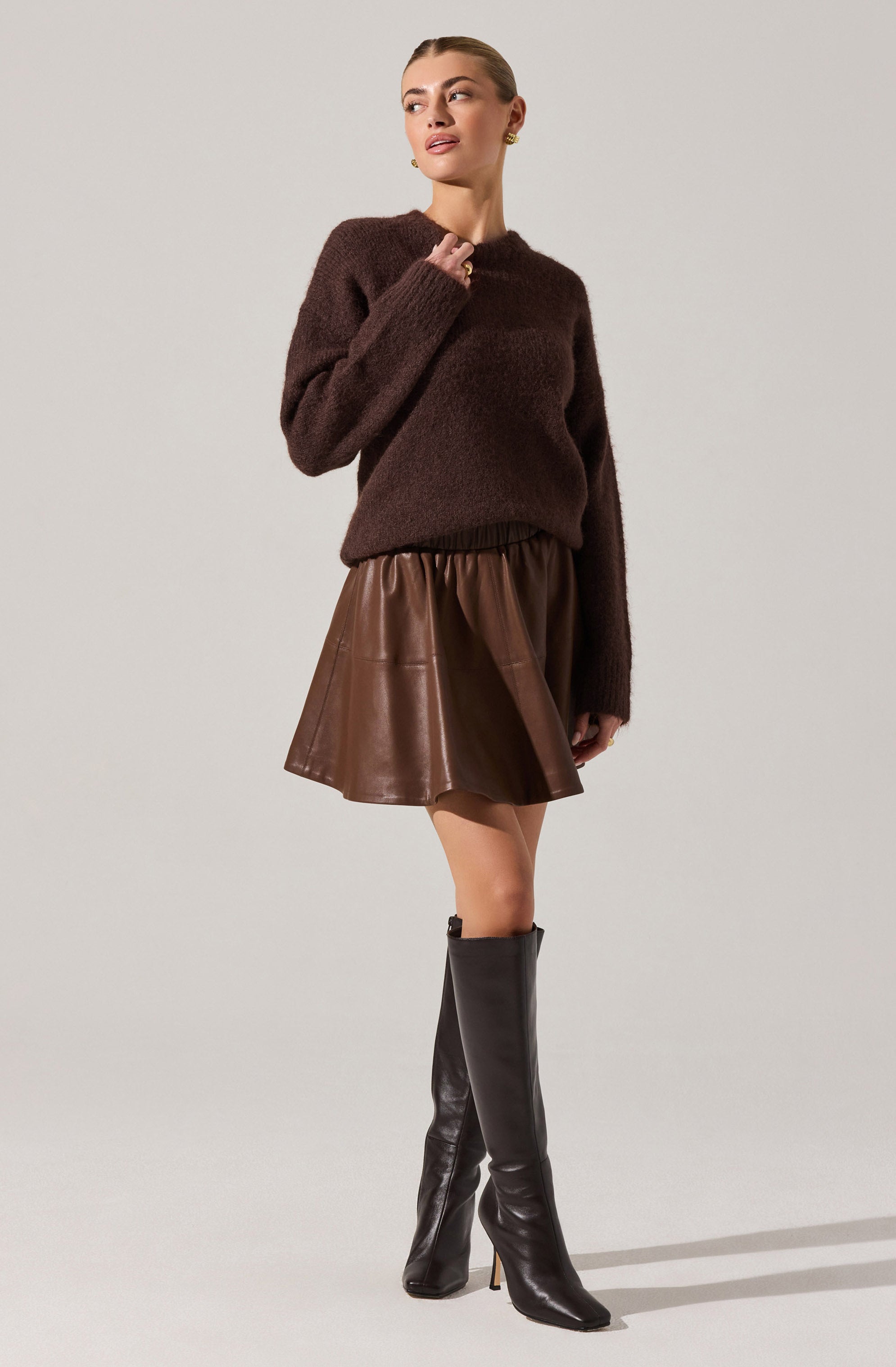 A woman is wearing a dark brown, fuzzy knit sweater with long sleeves and a rounded neckline. She pairs it with a high-waisted, brown leather skirt that has a flared silhouette. She completes the look with tall, black leather heeled boots that reach just below the knee.