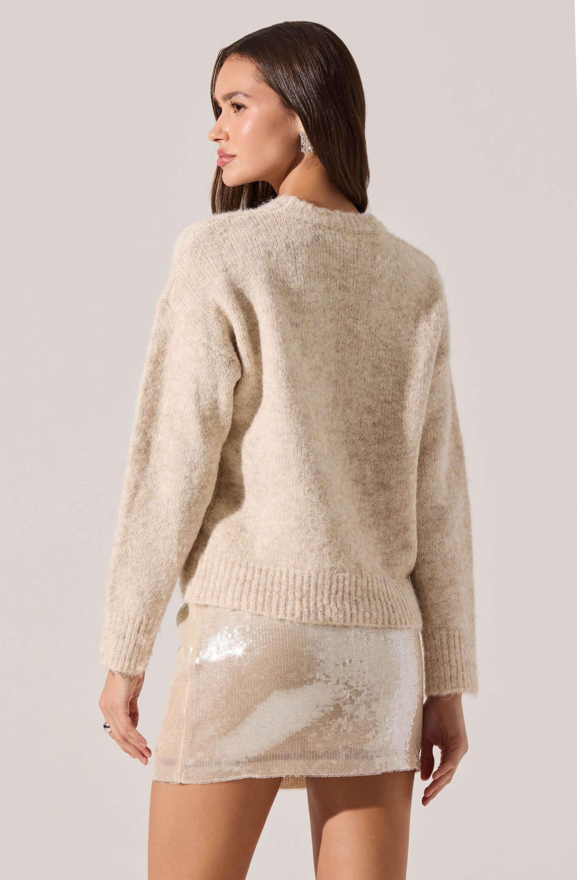 A woman is wearing a beige, fuzzy, long-sleeved sweater with a crew neckline and ribbed hem. She pairs it with a shiny, light-colored sequined mini skirt.