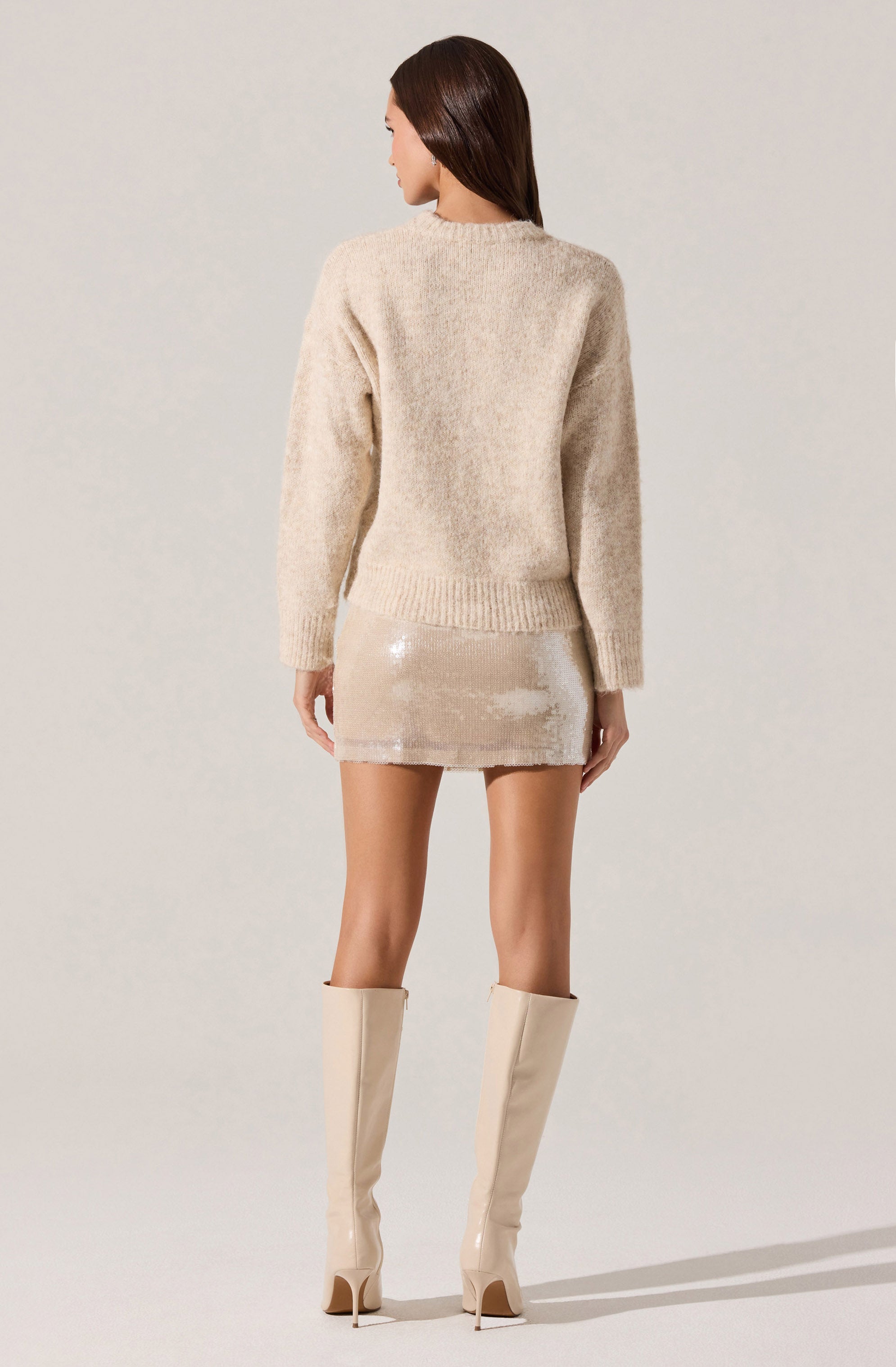 The woman is wearing a loose, long-sleeved, beige knitted sweater with a round neckline. She has on a short, shiny, beige sequin skirt and tall, matching beige thigh-high boots with high heels.