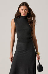 A woman is wearing a sleeveless, dark gray turtleneck top and a matching high-waisted, long skirt. She accessorizes with gold hoop earrings, a large gold bracelet, and a black handbag.