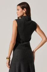 A woman is wearing a sleeveless, high-neck, dark gray knit top and a matching high-waisted skirt. The outfit appears elegant and fitted, emphasizing a sophisticated style. She accessorizes with large gold hoop earrings and a chunky gold bracelet.
