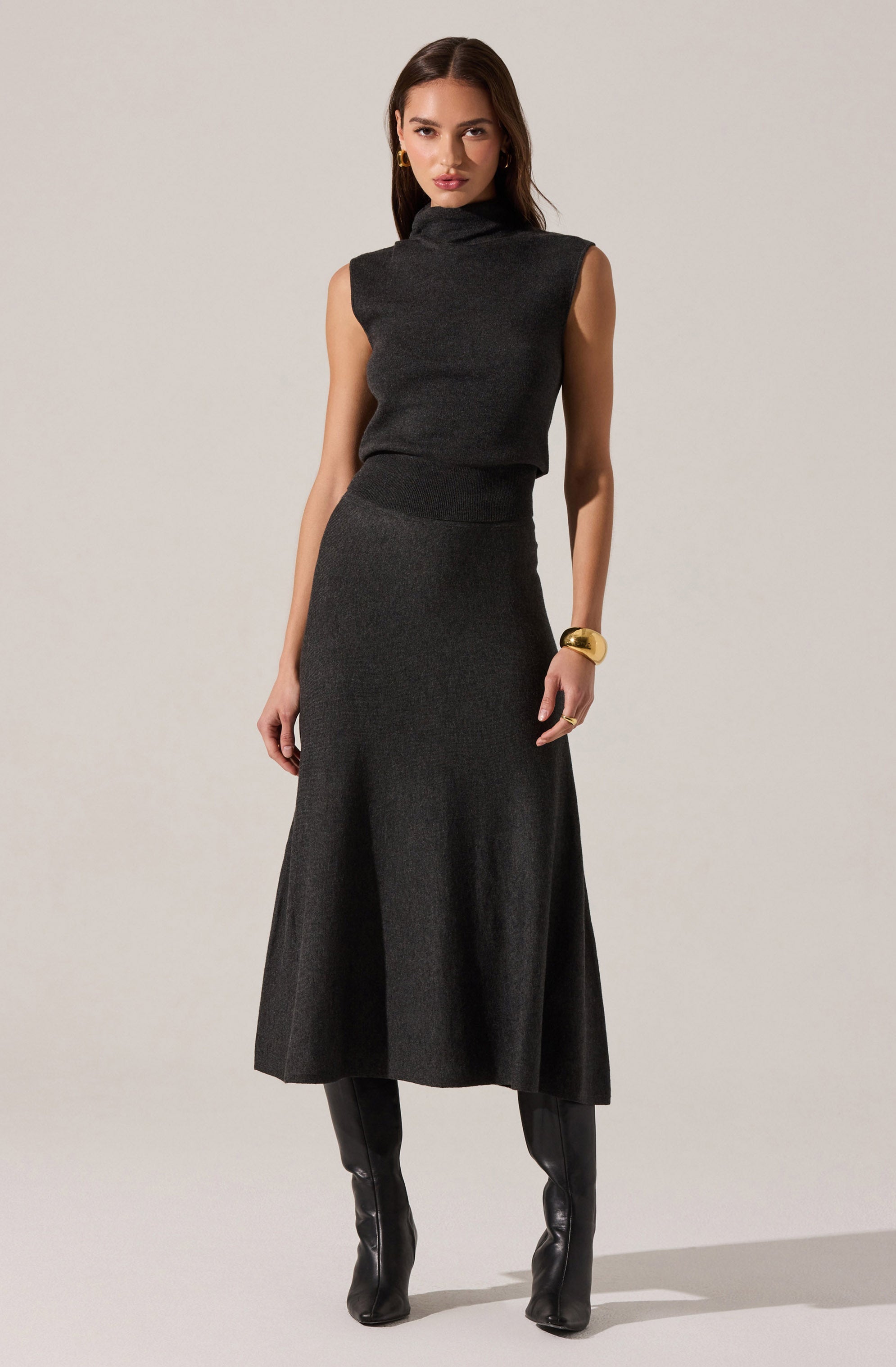 A woman is wearing a sleeveless, high-neck, dark gray top paired with a matching, high-waisted, long A-line skirt that falls to her ankles. She accessorizes with a large gold cuff bracelet on her right wrist and gold hoop earrings. She has on knee-high black leather boots.