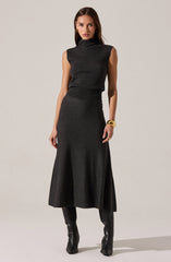 A woman is wearing a sleeveless, high-neck, dark gray top paired with a matching, high-waisted, long A-line skirt that falls to her ankles. She accessorizes with a large gold cuff bracelet on her right wrist and gold hoop earrings. She has on knee-high black leather boots.