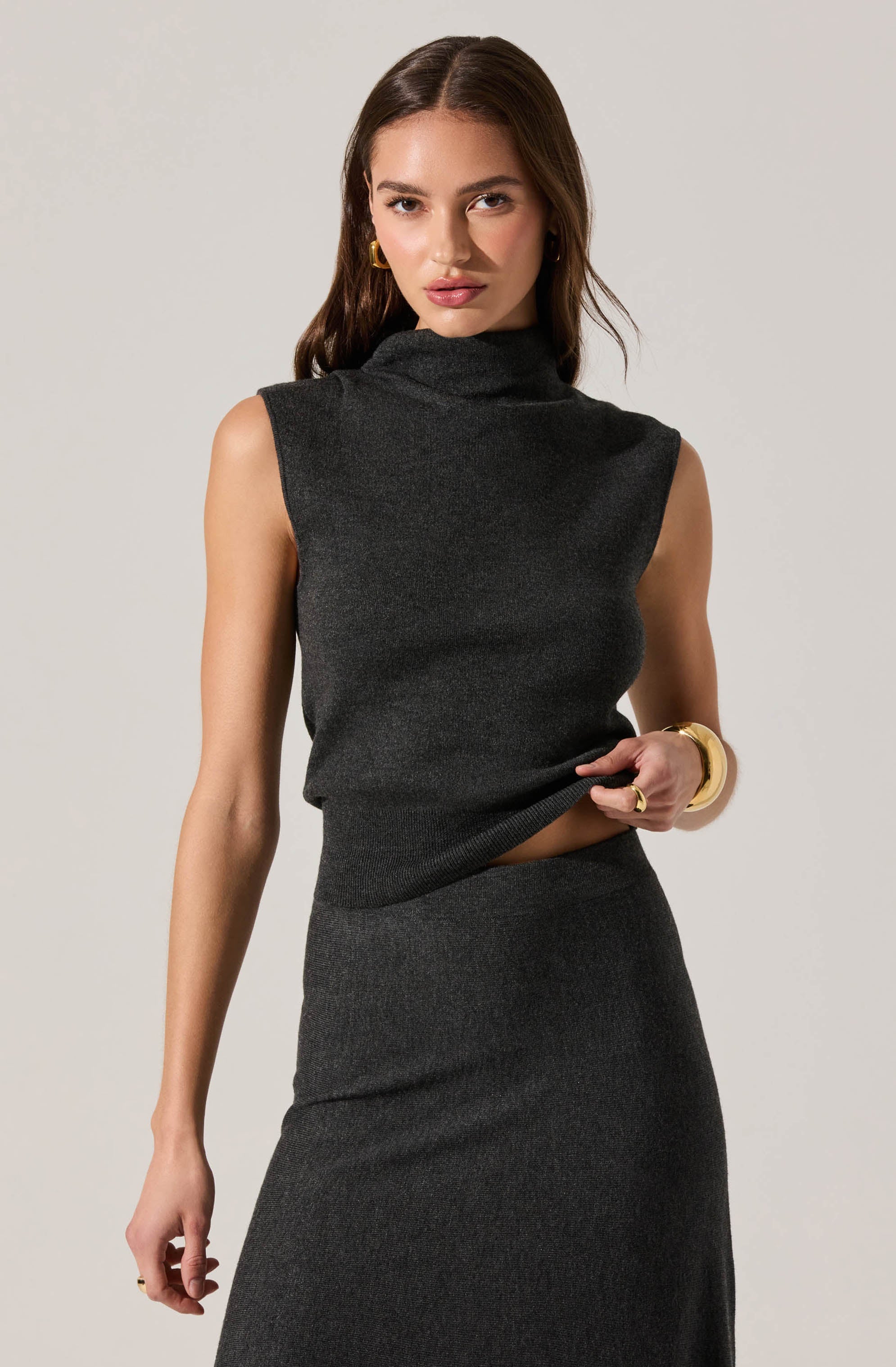 A woman is wearing a sleeveless, dark gray, turtleneck top with a loose fit, paired with a matching high-waisted skirt. She accessorizes with gold hoop earrings, a gold bracelet, and a ring. Her hair is styled in loose waves.