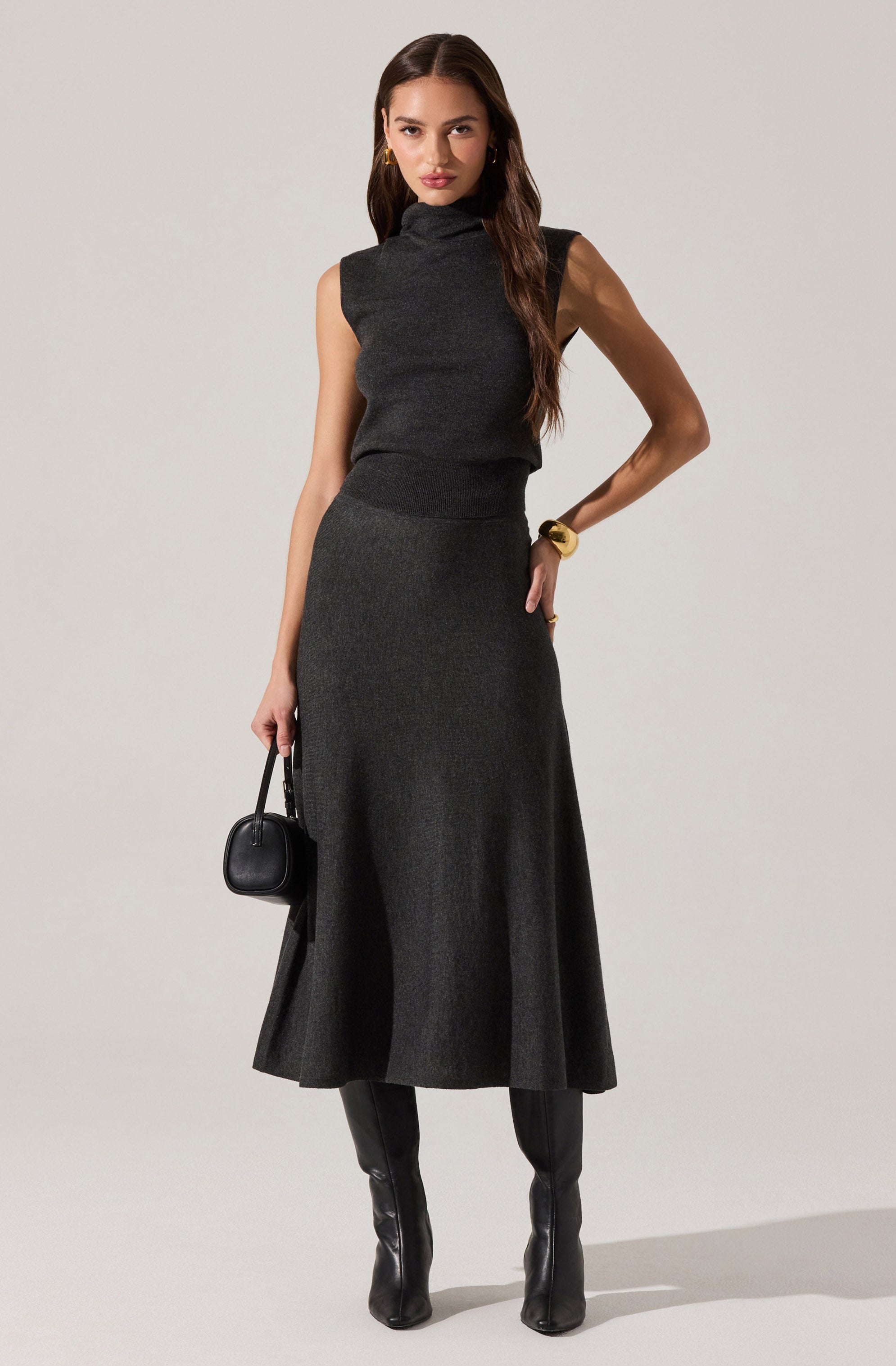 A woman is wearing a sleeveless, dark gray turtleneck top paired with a matching long, flared skirt. She has gold hoop earrings, a wide gold bracelet, and is holding a small black handbag. She is also wearing black knee-high boots.