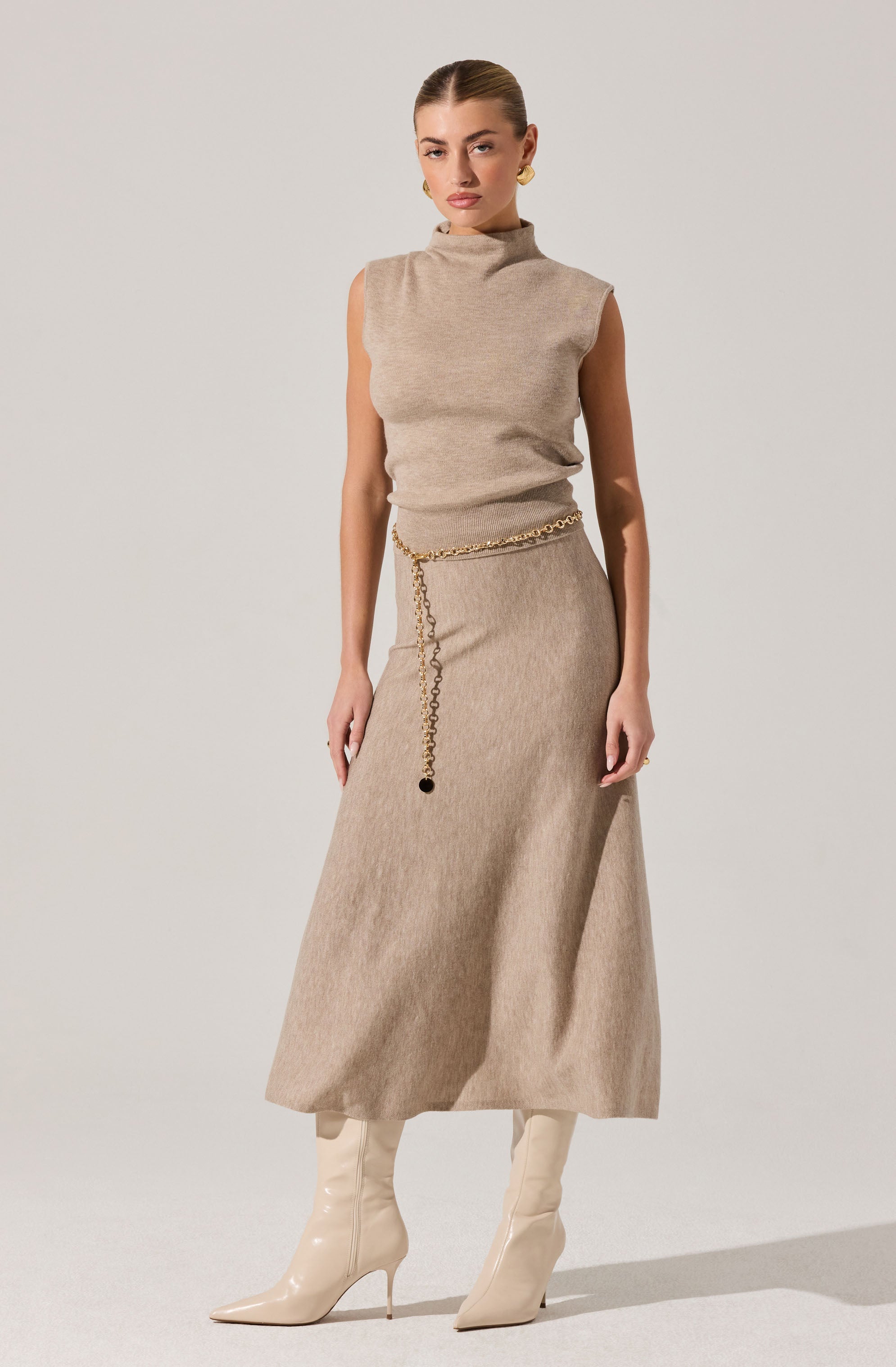 The woman is wearing a sleeveless, high-neck beige top made of a soft, knit fabric, paired with a matching long beige skirt that reaches above her ankles. She accessorizes with a gold chain belt around her waist and gold hoop earrings. She completes her outfit with off-white, shiny, knee-high heeled boots.