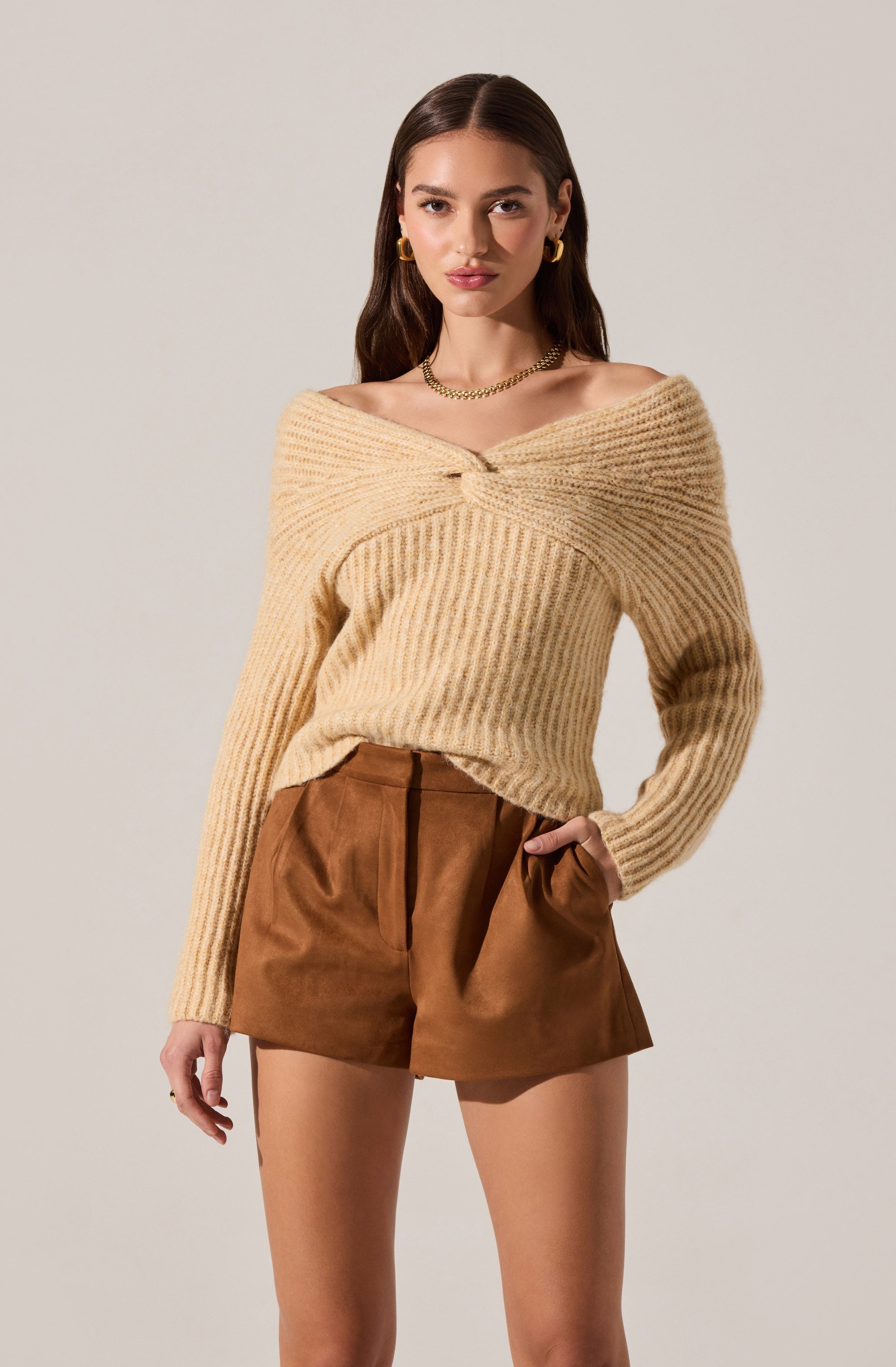 A woman is wearing a cream-colored, off-the-shoulder knit sweater with a twisted front detail, paired with high-waisted brown shorts. She accessorizes with gold hoop earrings and a gold chain necklace.