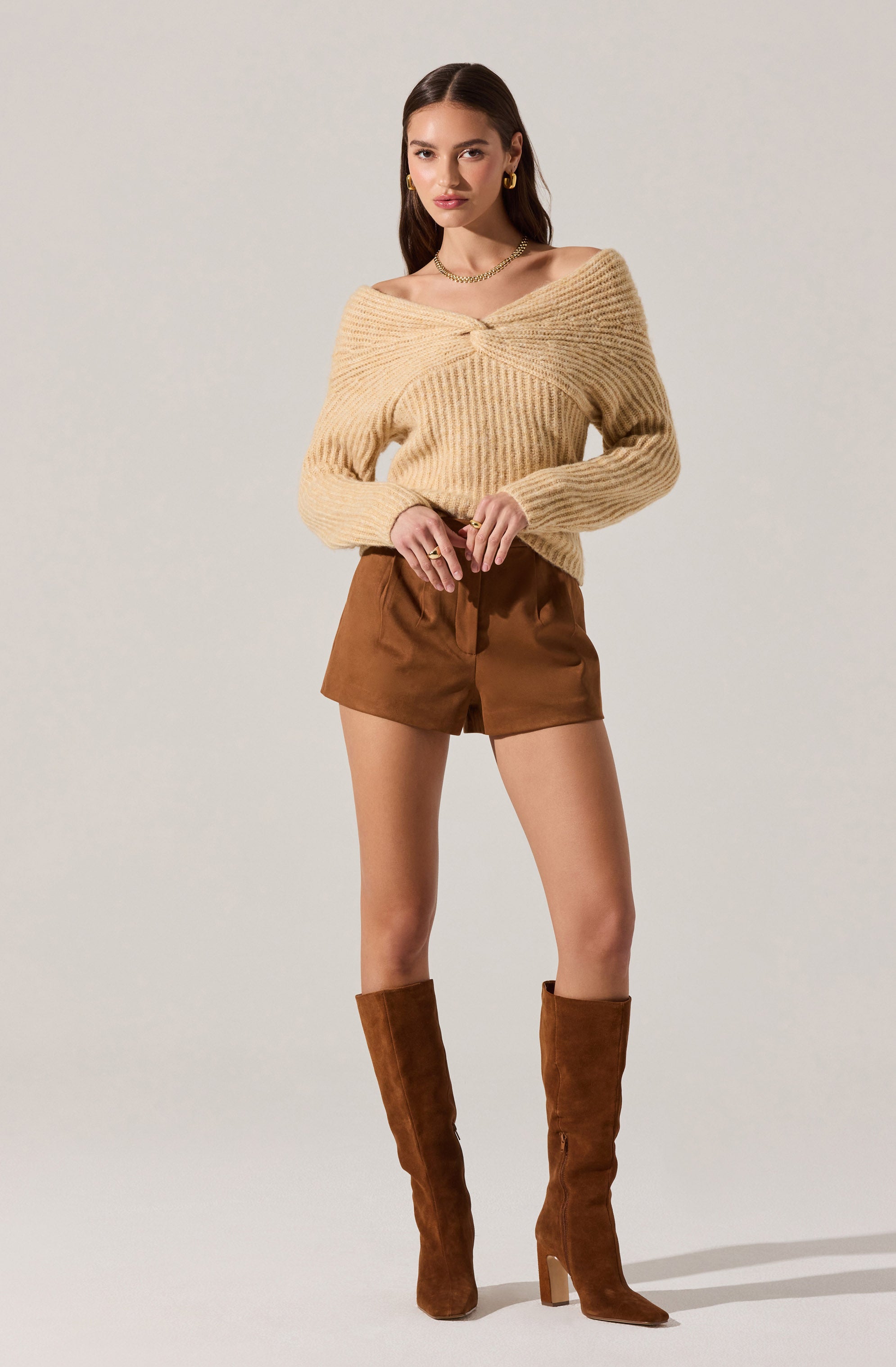 A woman is wearing an oversized, chunky knit sweater in a light beige color with a twisted knot detail at the neckline. She is also dressed in high-waisted, loose-fitting brown shorts. Her outfit is completed with tall, brown suede boots that reach just below her knees. She accessorizes with gold hoop earrings, a gold necklace, and rings.