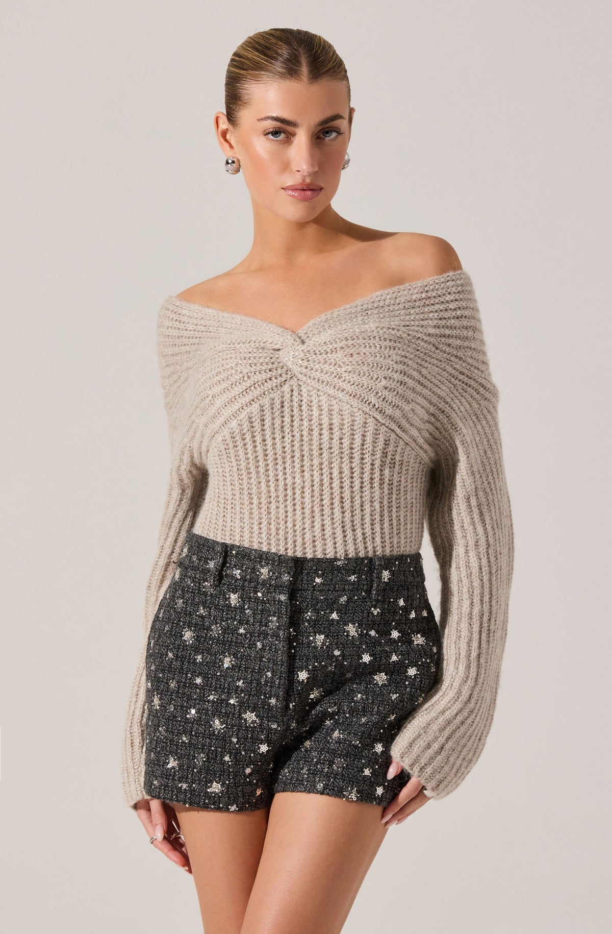 A woman is wearing a beige, off-the-shoulder knit sweater with a twisted front detail and long sleeves. She is also wearing high-waisted, black textured shorts with a white floral pattern.