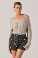 A woman is wearing a beige, off-the-shoulder knit sweater with a twisted front detail and long sleeves. She is also wearing high-waisted, black textured shorts with a white floral pattern.