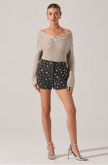 A woman is wearing a beige, chunky-knit sweater with a twisted, off-the-shoulder neckline. She pairs it with high-waisted black shorts decorated with a white star pattern. She completes her outfit with silver, pointed-toe high heels that have ankle straps.