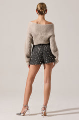 A woman is wearing an oversized, chunky beige knit sweater with a wide neckline that reveals her shoulders. She pairs the sweater with high-waisted black shorts that are decorated with a pattern of white and silver star-shaped embellishments. She completes the outfit with elegant silver and white high-heeled sandals with ankle straps. Her hair is styled in a neat, low bun.