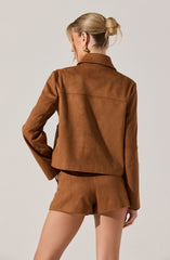 A woman is wearing a brown suede jacket and matching brown suede shorts. The jacket has a collar and long sleeves, and the shorts are fitted with a simple waistband. The outfit appears stylish and coordinated in tone and material.