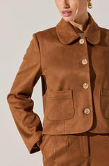 A woman is wearing a brown, tailored jacket made of a faux suede material. The jacket features a rounded collar, four large buttons down the front, and two patch pockets on each side at the front. The sleeves are long, and she has one hand in her pocket.