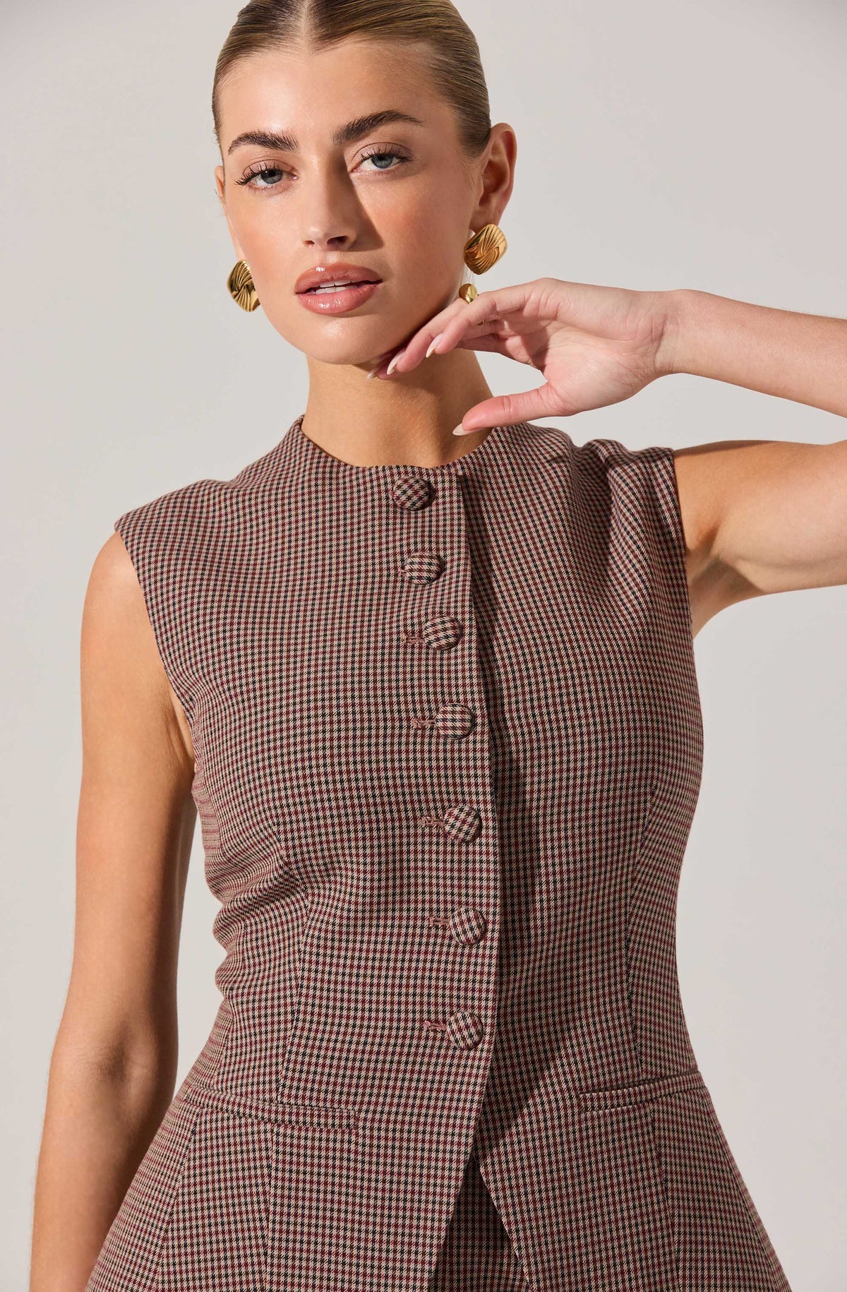A woman is wearing a sleeveless, fitted dress with a high neckline and a small checkered pattern in shades of brown and black. The dress features a row of fabric-covered buttons down the front and two small pockets on the lower front. She is accessorized with large, gold, geometric earrings.