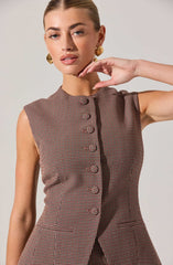 A woman is wearing a sleeveless, fitted dress with a high neckline and a small checkered pattern in shades of brown and black. The dress features a row of fabric-covered buttons down the front and two small pockets on the lower front. She is accessorized with large, gold, geometric earrings.