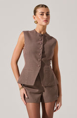 The woman is wearing a sleeveless, tailored vest with a high neckline and a row of buttons down the front, made of fabric with a small checkered pattern in shades of brown and black. She pairs it with matching high-waisted shorts that have the same checkered pattern.