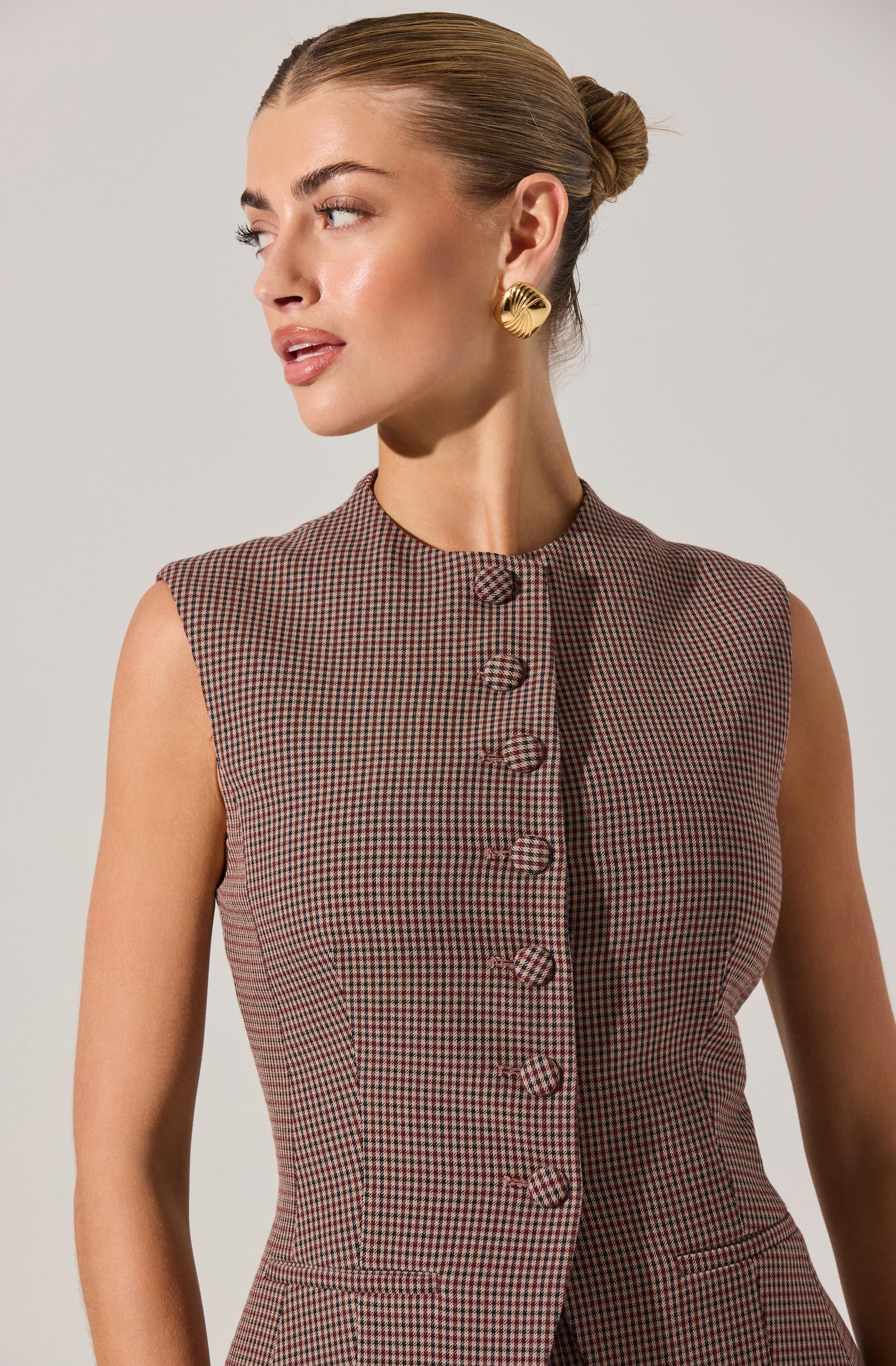 A woman is wearing a sleeveless, fitted dress with a high, round neckline. The dress features a small checkered pattern in shades of brown and red, with a row of fabric-covered buttons down the front. She has her hair styled in a sleek bun and is accessorized with large, gold, shell-shaped earrings.