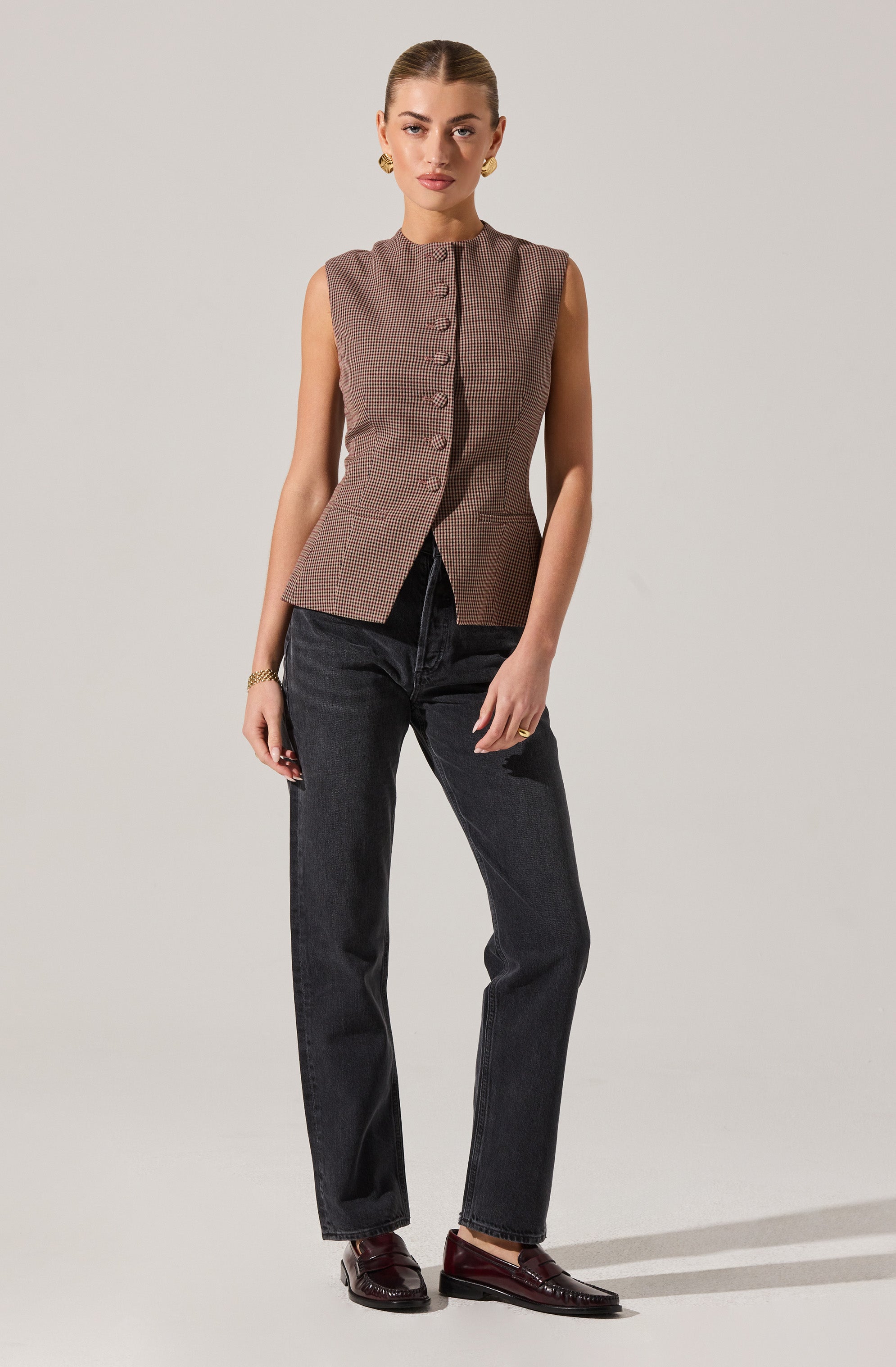 A woman is wearing a sleeveless, button-up vest with a small, checkered pattern in shades of brown and beige. She pairs it with high-waisted, dark-wash jeans and dark brown loafers. She accessorizes with gold jewelry, including earrings, a bracelet, and rings.