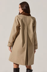 A woman is wearing a beige trench coat with a brown collar. The coat has long sleeves with decorative buttons near the cuffs, and it features a pleat at the back for movement. The coat is knee-length and appears to be made of a classic, smooth fabric.