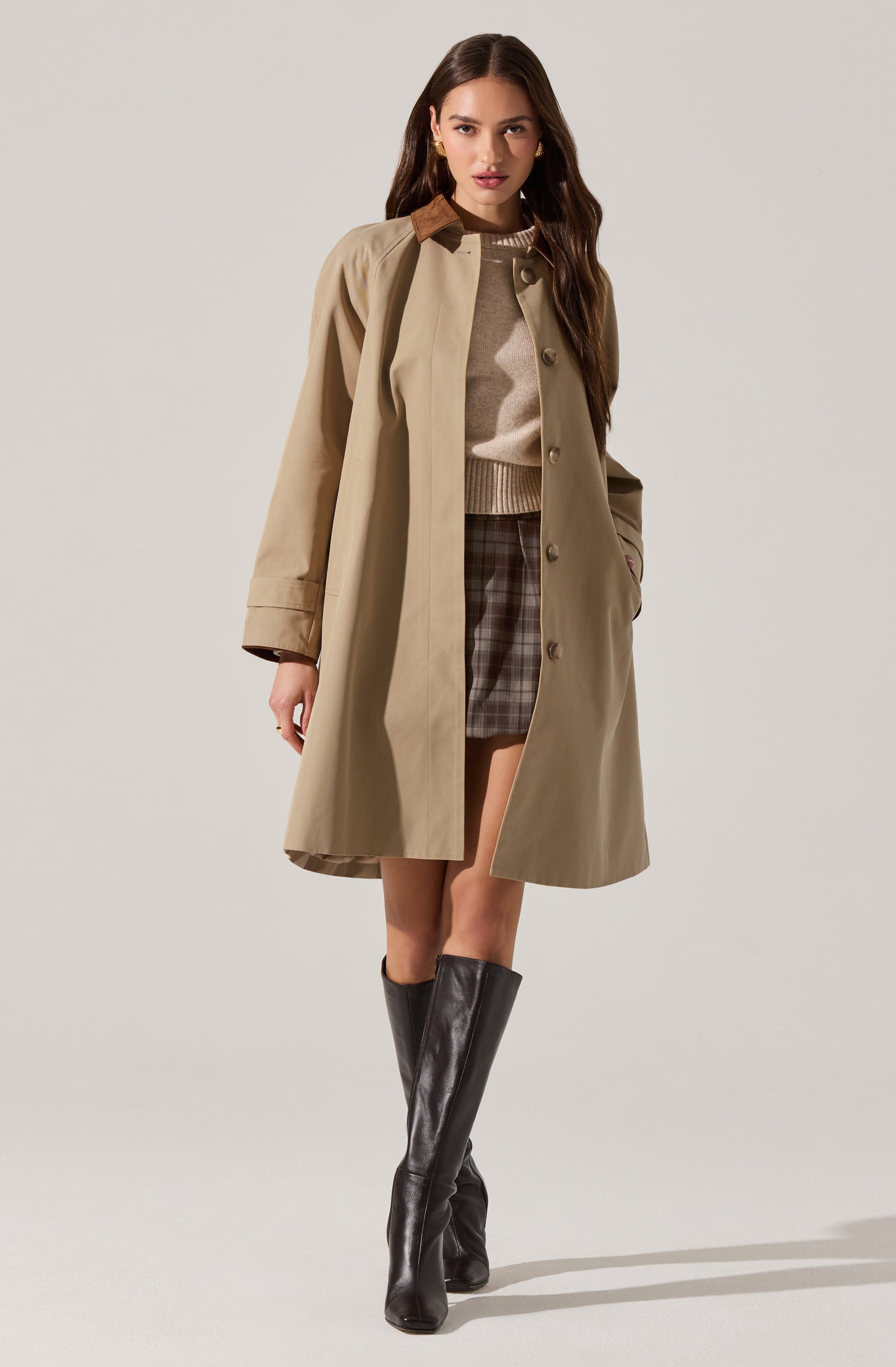A woman is wearing a beige trench coat with a brown collar, buttoned down the front. Underneath, she has on a light beige sweater with a ribbed hem. She pairs this with a plaid skirt in shades of brown and gray. She is also wearing knee-high black leather boots.