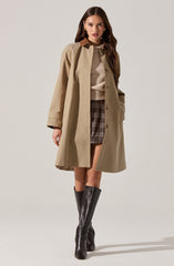 A woman is wearing a beige trench coat with a brown collar, buttoned down the front. Underneath, she has on a light beige sweater with a ribbed hem. She pairs this with a plaid skirt in shades of brown and gray. She is also wearing knee-high black leather boots.