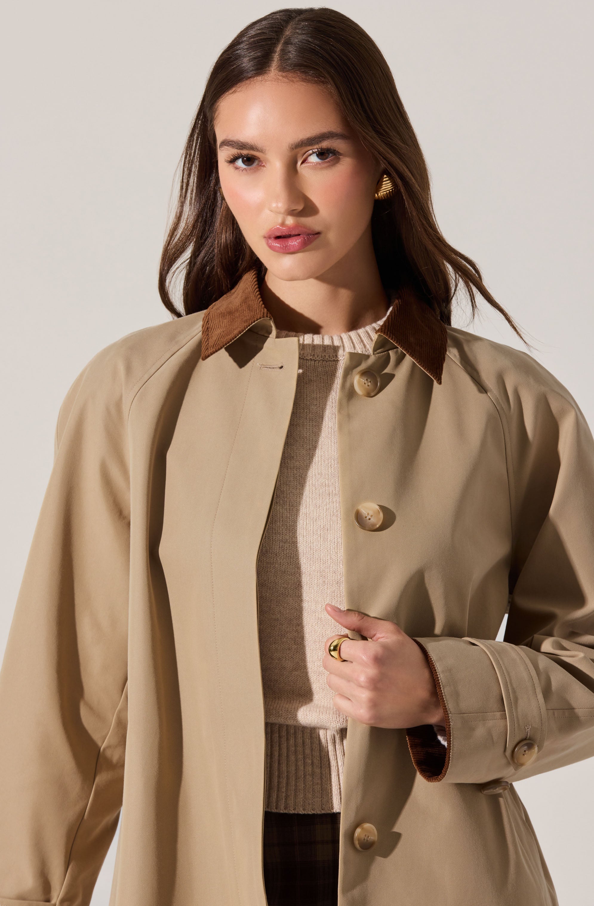 A woman is wearing a beige trench coat with a brown corduroy collar, beige buttons down the front, and a belt at the waist. Underneath, she has on a light-colored sweater.