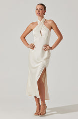 A woman is wearing a elegant, satin ivory dress with a halter neckline, featuring a twisted fabric detail at the neck and a keyhole cutout. The dress has a high slit on one side revealing her leg, and she is wearing gold strappy high-heeled sandals. She accessorizes with large hoop earrings and gold rings.