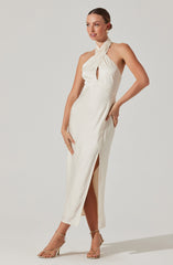 A woman is wearing an elegant, cream-colored satin dress with a halter neckline that features a twisted strap design and a small front keyhole cutout. The dress has a slit on the side, revealing her leg. She is accessorized with gold hoop earrings, a gold ring, and strappy high-heeled sandals.