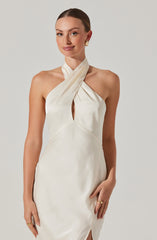 A woman wearing a sleeveless, elegant, white satin dress with a halter neck and a keyhole opening in the front. The dress has pleated fabric at the neck that wraps around her neck and drapes over her shoulder. She is accessorized with gold hoop earrings and gold rings.