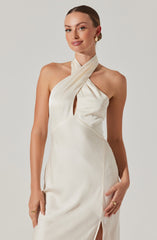 A woman wearing an elegant, sleeveless, pearl-colored satin dress with a halter neckline. The dress features a twisted, gathered detail at the neck with a keyhole cutout in the front, and a slit on the side. She accessorizes with gold hoop earrings and a gold ring.