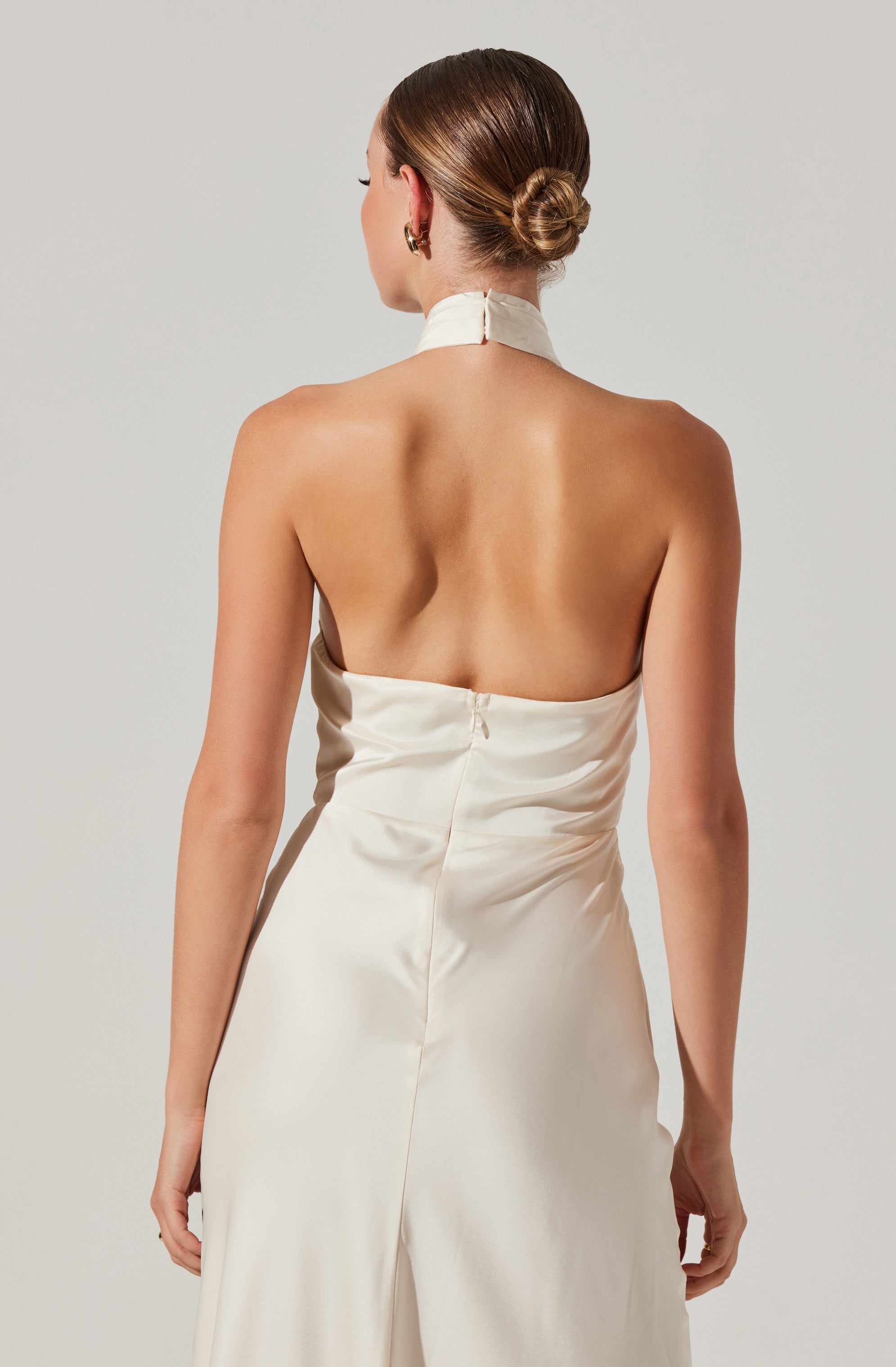 A woman wearing a sleeveless, backless, ivory satin dress with a high neck and a zipper at the back. Her hair is styled in a sleek, low bun, and she has gold hoop earrings.