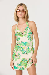 A woman is wearing a sleeveless, halter-neck dress with a colorful tropical floral pattern in shades of green, pink, and blue on a light cream background. The dress has a fitted bodice and a slightly loose, short skirt.