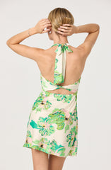 A woman is wearing a sleeveless, halter-neck dress with a floral print in shades of green, pink, and cream. The dress features a keyhole cutout at the back and a fabric tie at the neck. The overall look is light and summery with a colorful, tropical pattern.