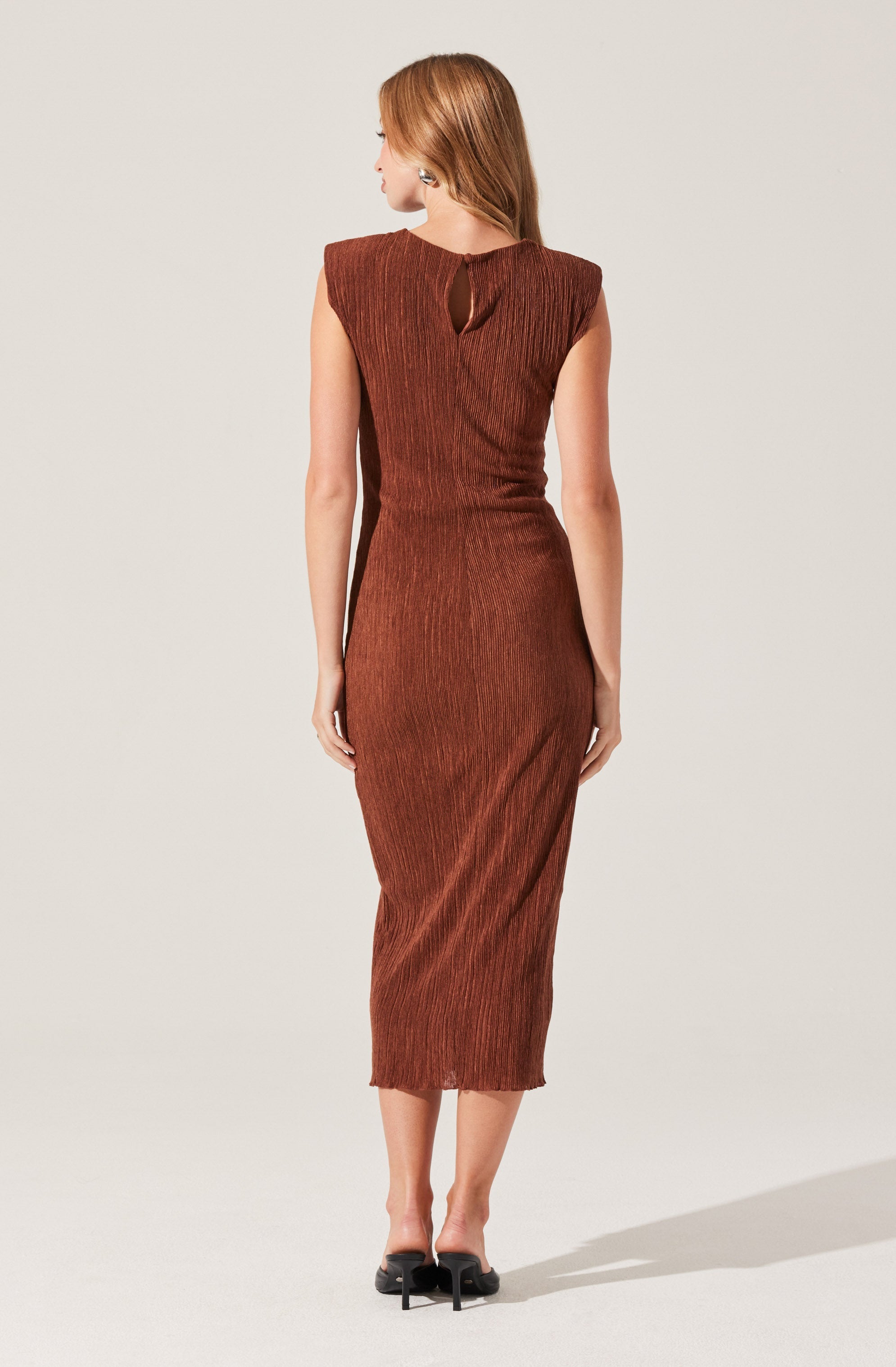 A woman is wearing a sleeveless, fitted midi dress in a rich, textured rust color. The dress features subtle vertical ribbing and a keyhole cutout at the upper back, near the neckline. She completes her look with black high-heeled shoes.