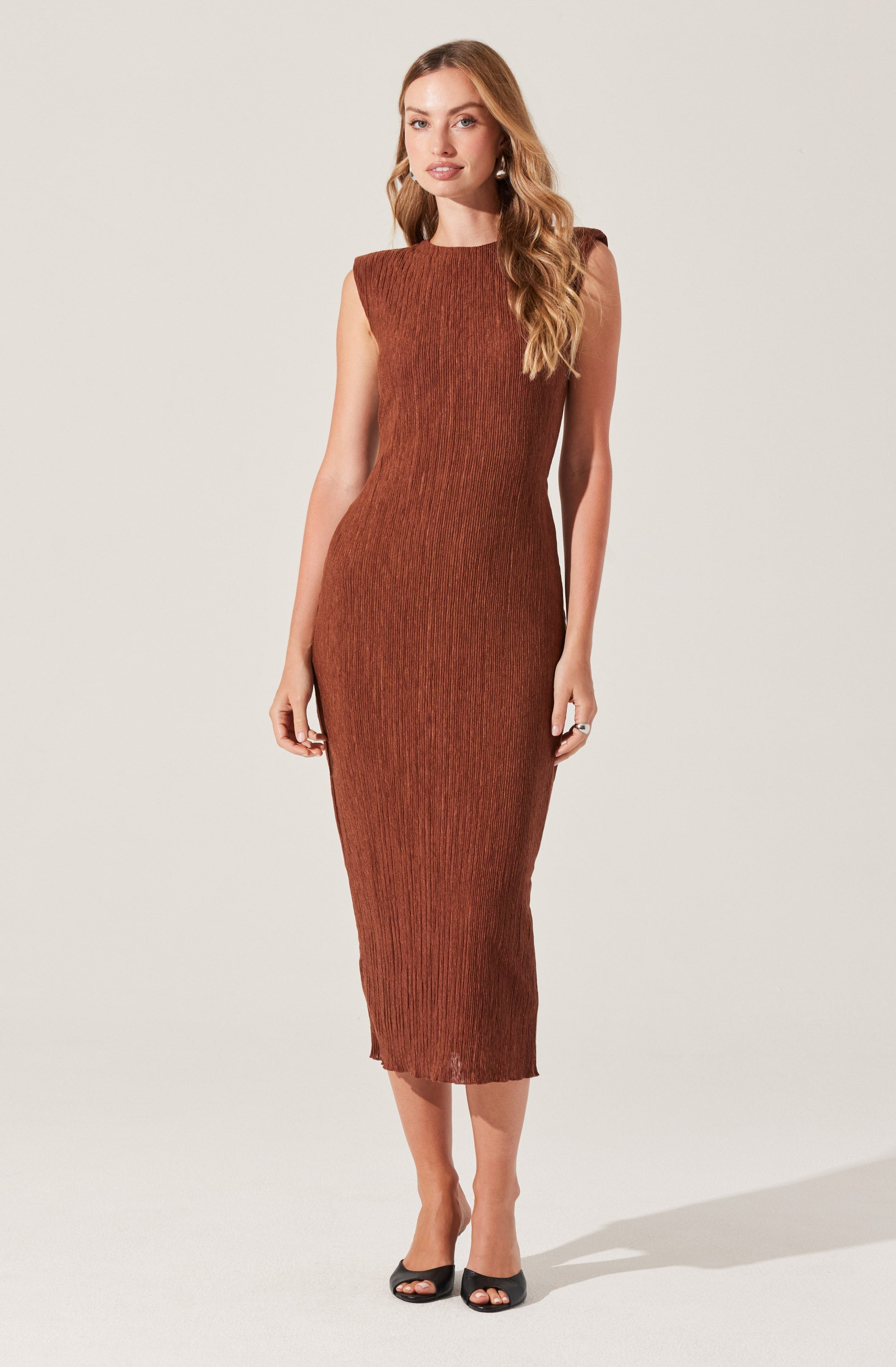 A woman is wearing a sleeveless, form-fitting, knee-length dress in a rust color with a textured, pleated pattern. She has long, wavy blonde hair and is wearing black open-toe heels.