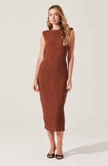 A woman is wearing a sleeveless, form-fitting, knee-length dress in a rust color with a textured, pleated pattern. She has long, wavy blonde hair and is wearing black open-toe heels.