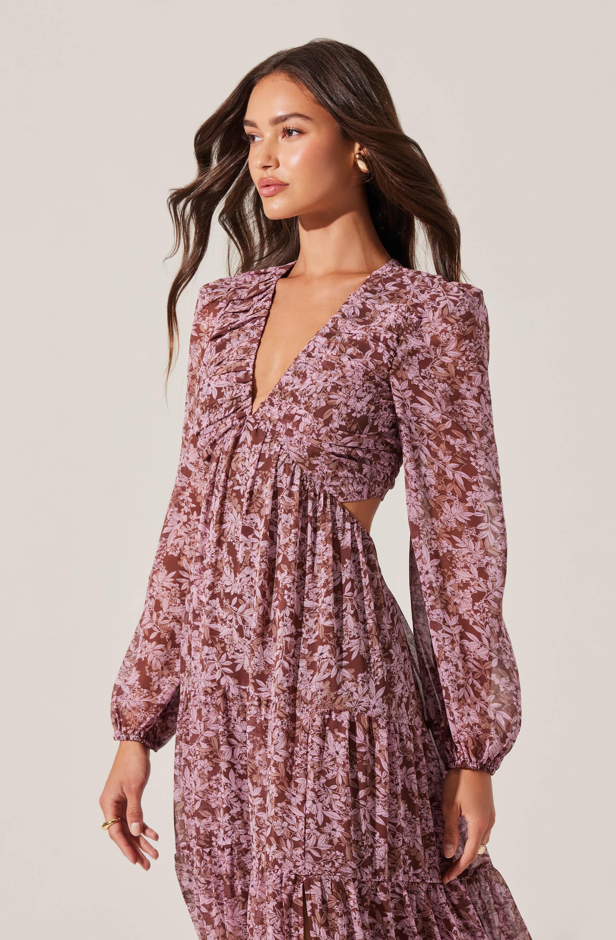 A woman is wearing a long, flowy dress with a pink and brown floral pattern. The dress features long, slightly puffed sleeves with elastic cuffs, a deep V neckline, and a fitted waist that creates a gathered, ruched effect across the bust. The dress has cut-outs at the sides, exposing skin on the waist, and the skirt is tiered with ruffled layers.