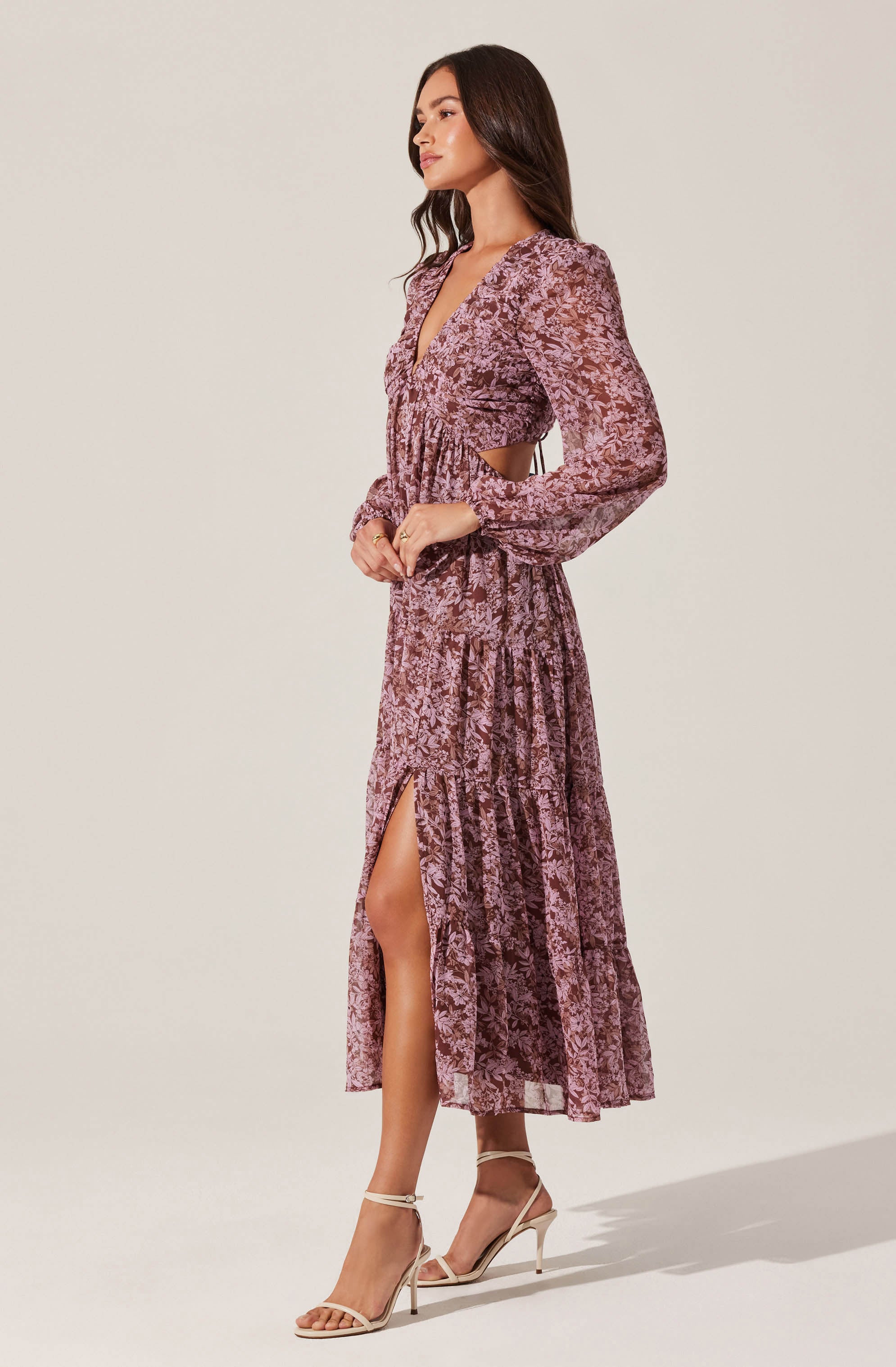 A woman is wearing a long, flowy dress with a mauve and pink floral pattern. The dress features a deep V-neck, long sheer sleeves, and an elastic waistband. It has a high slit on one side, revealing her leg, and cutouts at the waist. She is also wearing strappy, nude high-heeled sandals.