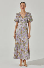 A woman is wearing a long, floral-patterned dress with a V-neckline. The dress features short, puffed sleeves and a side slit that extends up to her thigh. The fabric is decorated with a multicolored floral print in shades of purple, green, orange, and beige.
