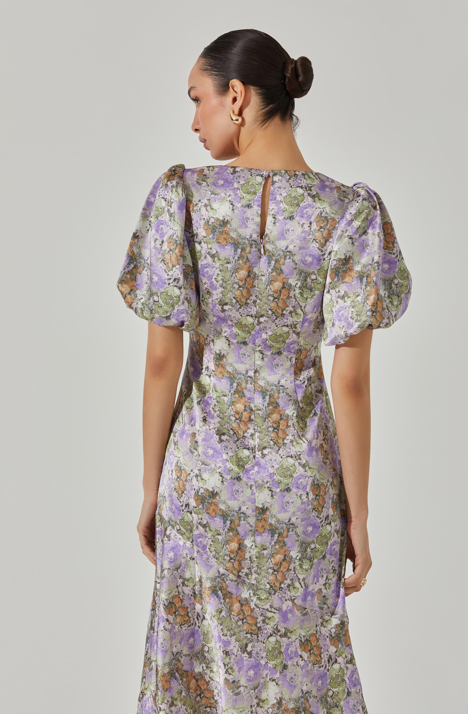 A woman is wearing a long dress with a floral pattern in shades of purple, green, orange, and cream. The dress features puffed short sleeves and a small keyhole opening at the back near the neckline.