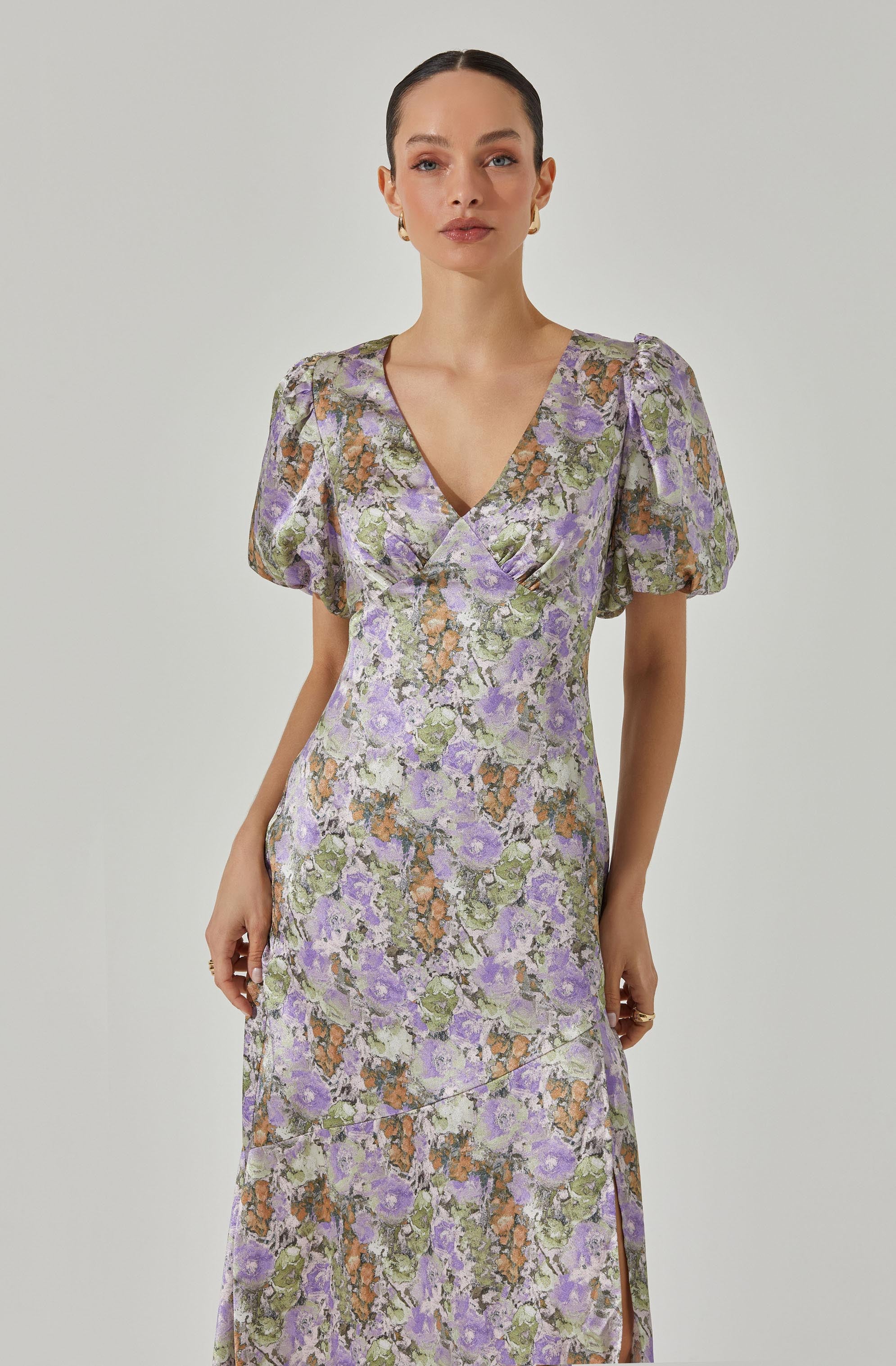 A woman is wearing a long dress with a floral pattern in shades of purple, green, orange, and cream. The dress has short, puffed sleeves with slight gathers at the shoulders, a V-neckline, and a fitted waist. The fabric appears to have a textured or slightly shiny surface.