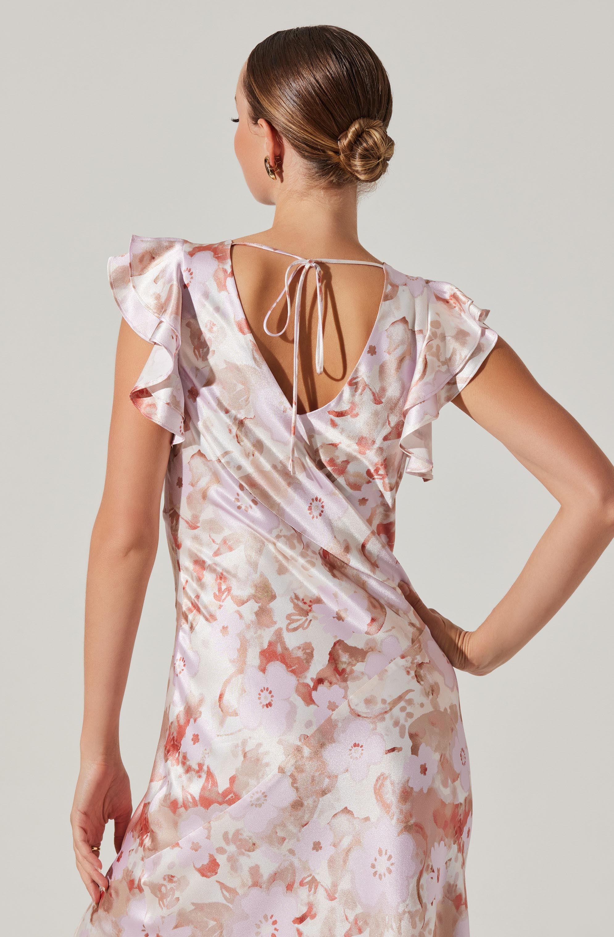 A woman is wearing a satin dress with a floral pattern in shades of pink, peach, and cream. The dress features ruffled cap sleeves and a deep V neckline at the back, tied with a delicate string.