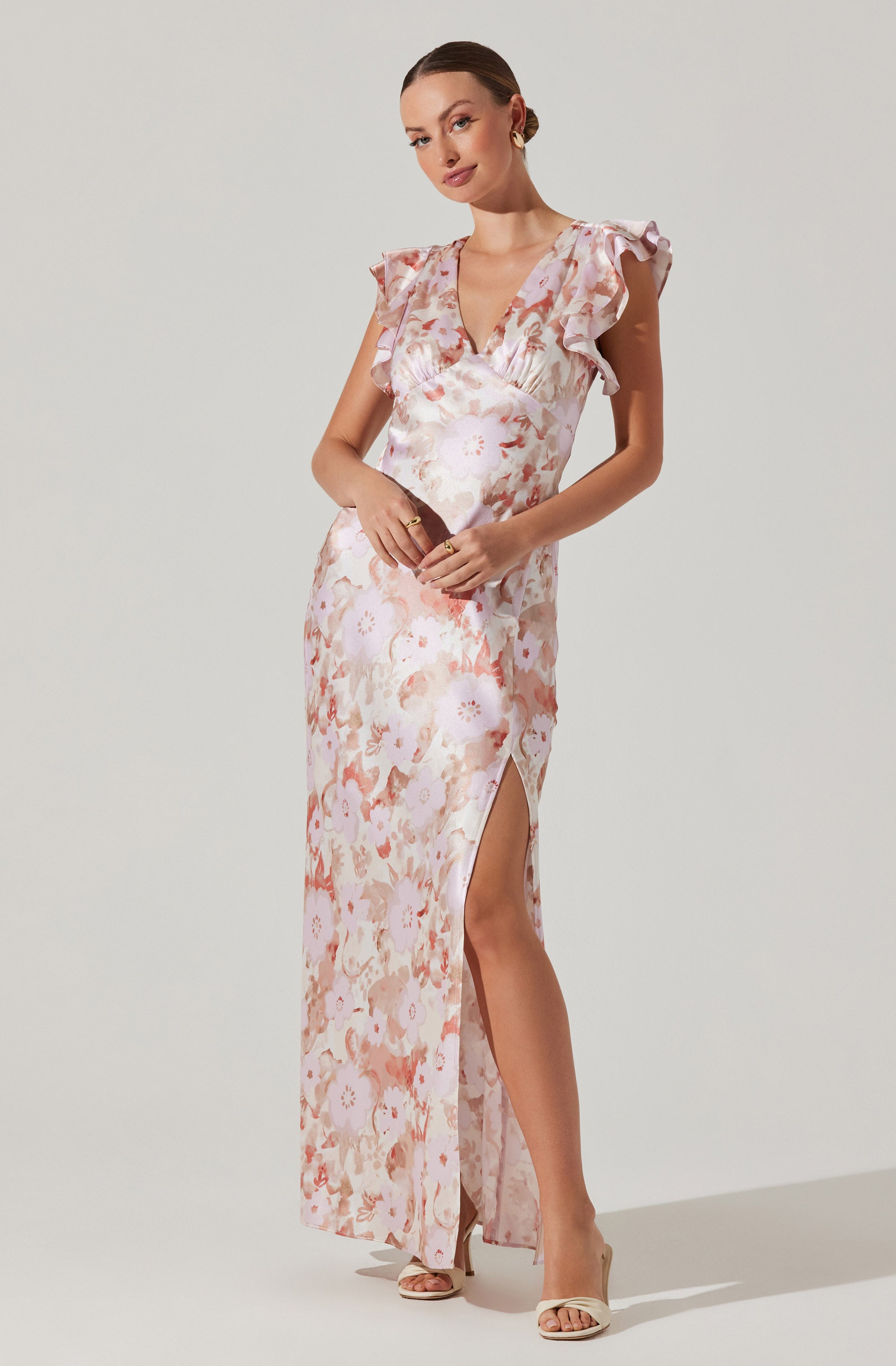 A woman is wearing a sleeveless, floor-length dress with a floral pattern in soft pastel shades of pink, peach, and white. The dress features ruffled cap sleeves, a V-neckline, and a high slit on one side, revealing her leg. She is paired with nude high-heeled sandals.